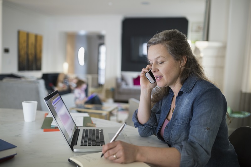 A woman speaking on the phone while sitting in front of a laptop and taking notes